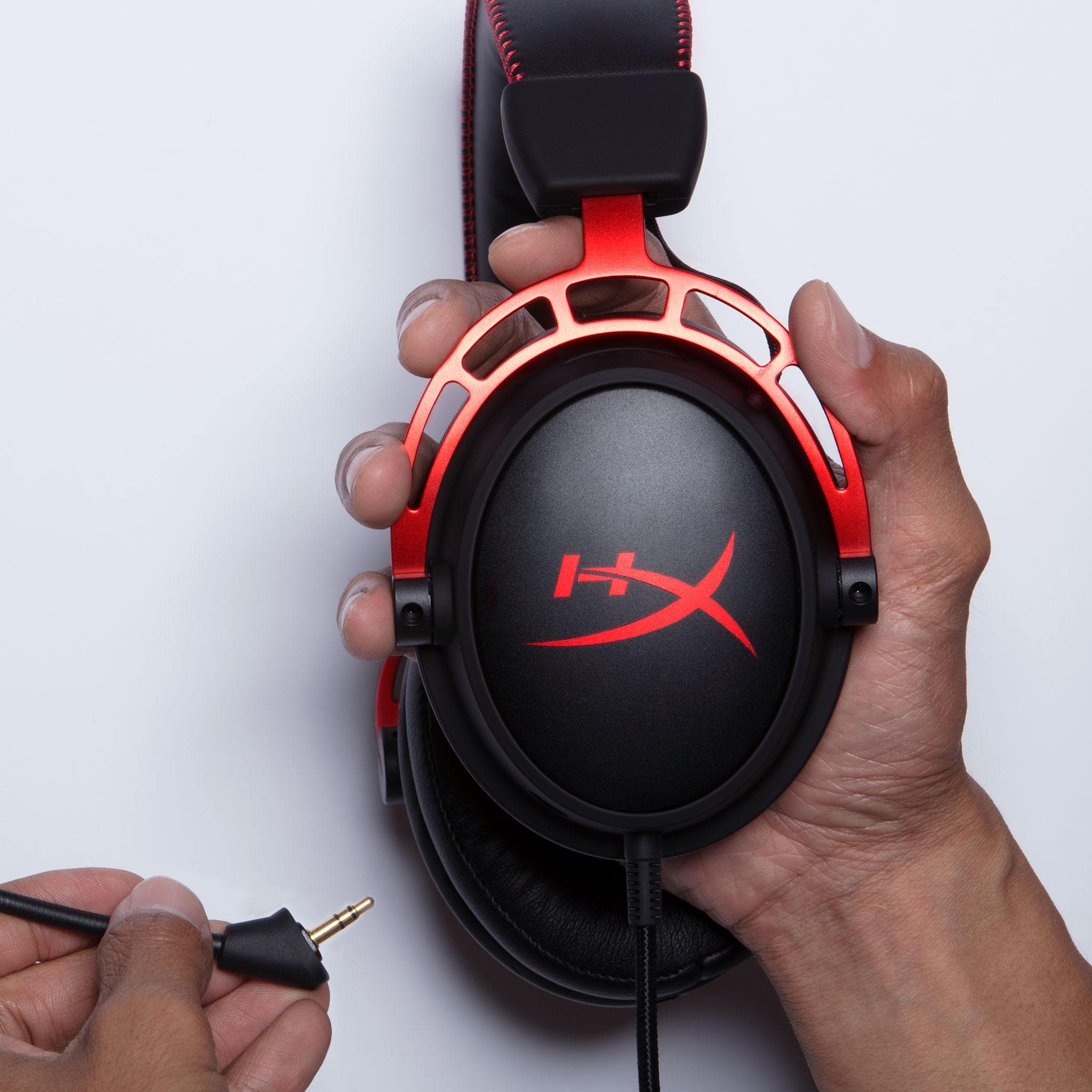 Hands holding a red and black HyperX Cloud Alpha gaming headset with detachable audio jack