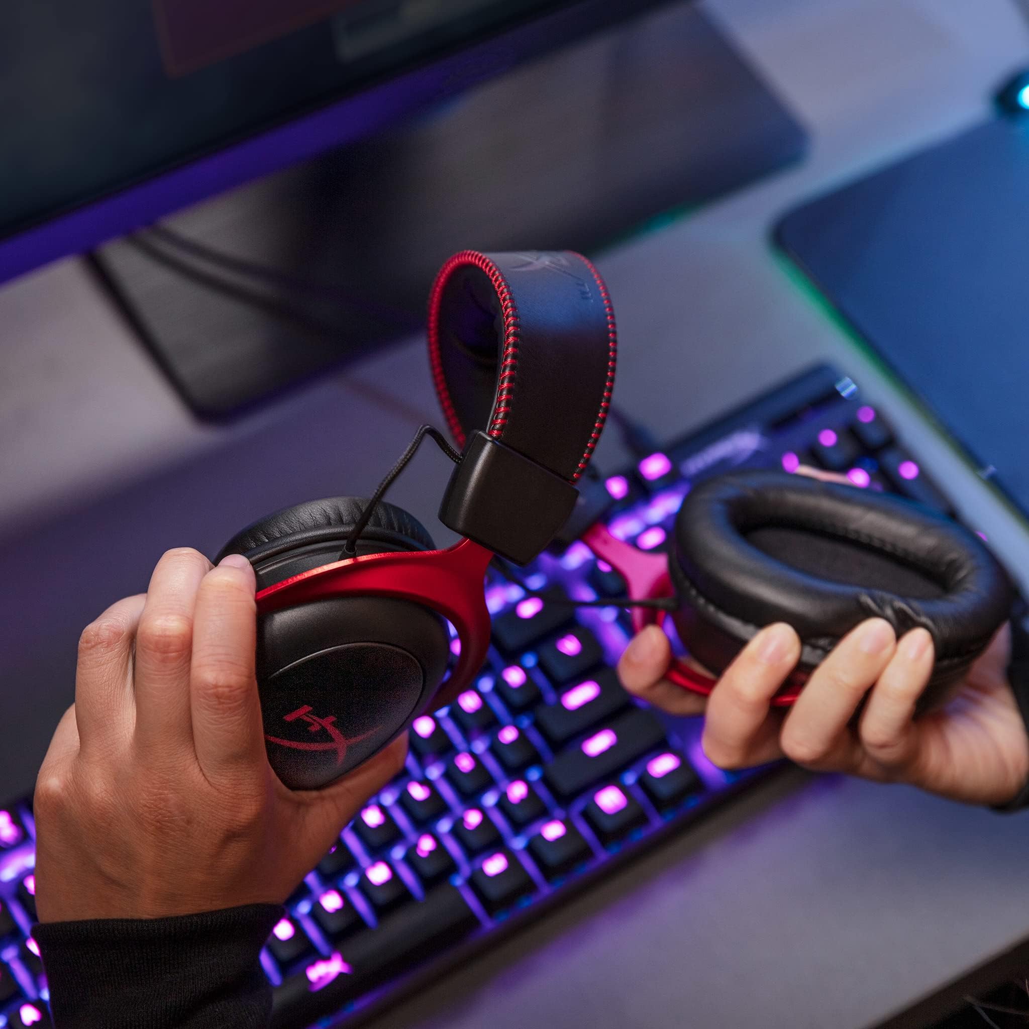 Hands holding red and black HyperX Cloud II gaming headset over a colorful backlit keyboard