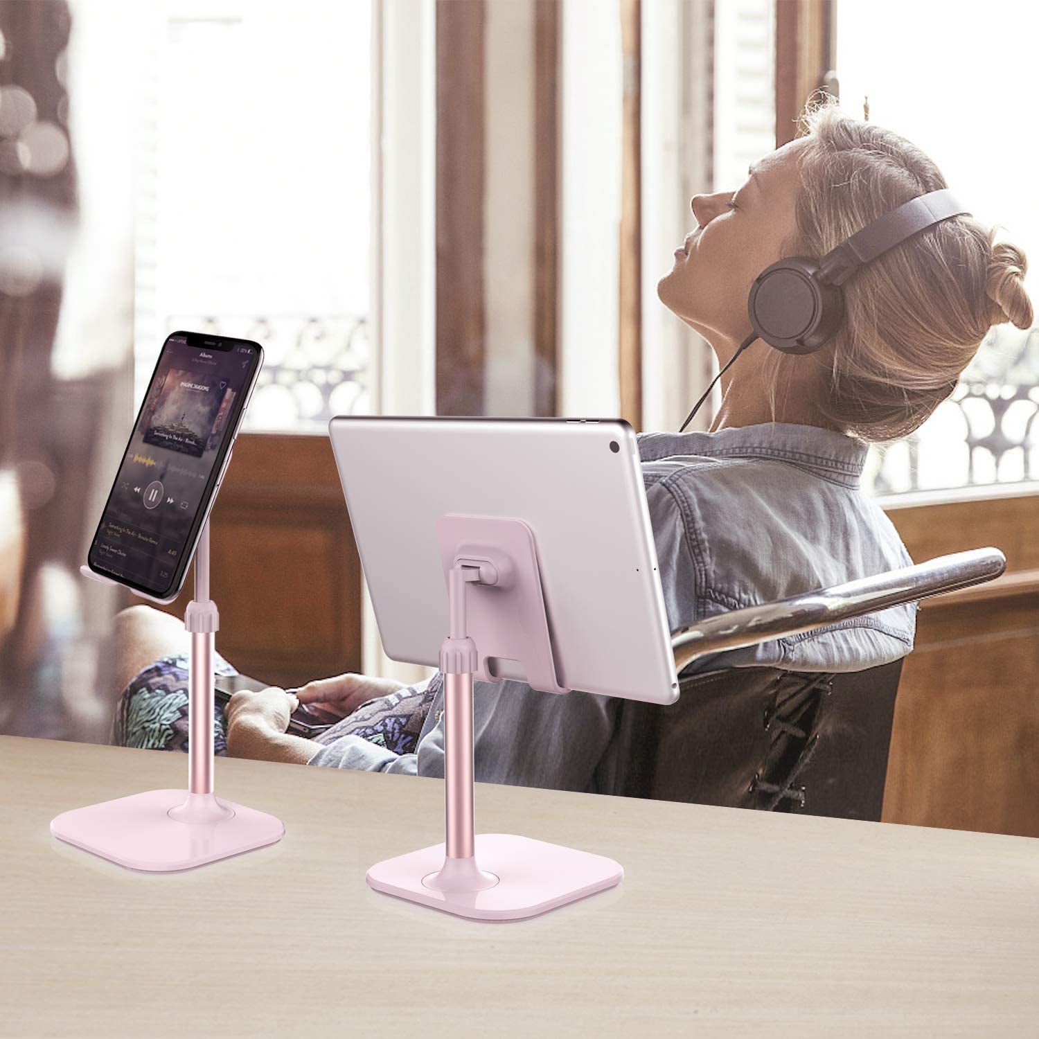 Pink adjustable cell phone stand for desk holding a phone and tablet with woman wearing headphones in background
