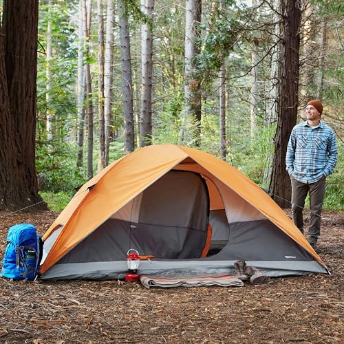 Orange and gray dome camping tent with rainfly set up in a forest with camping gear and a man nearby