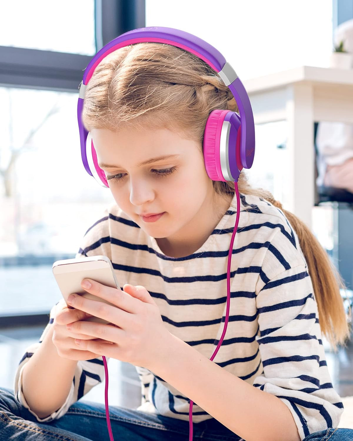 Young girl wearing purple and pink kids foldable headphones while using a smartphone indoors