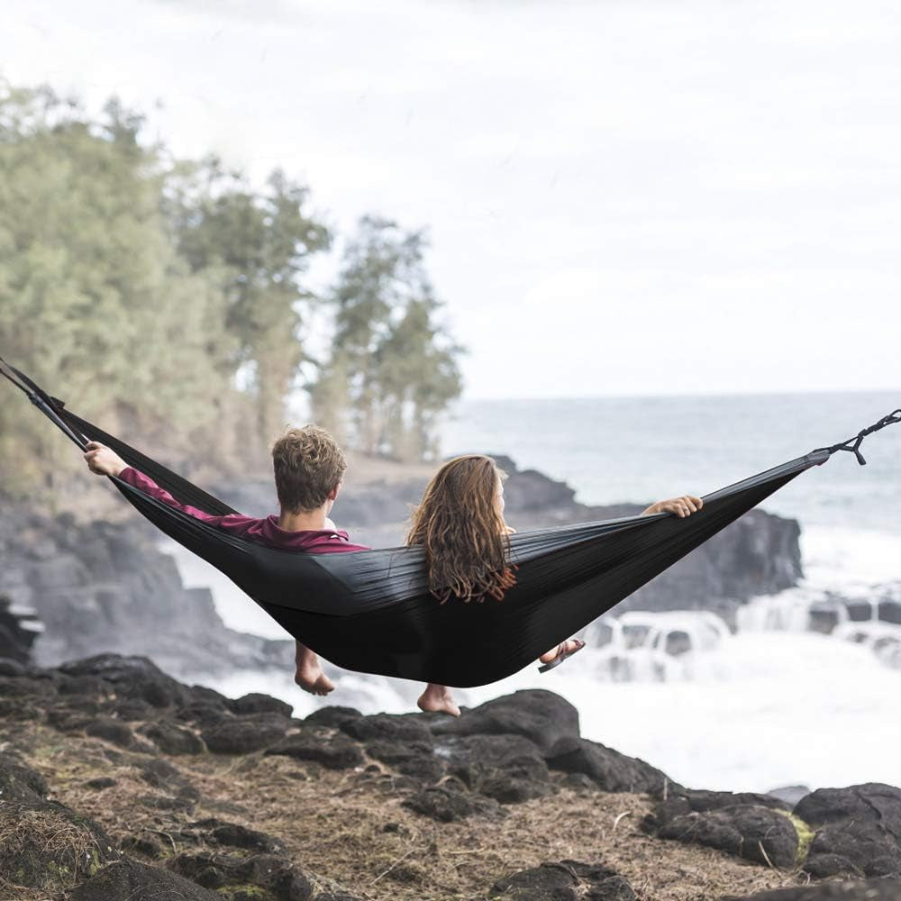 Couple relaxing in black double camping hammock with straps near rocky coastline and ocean