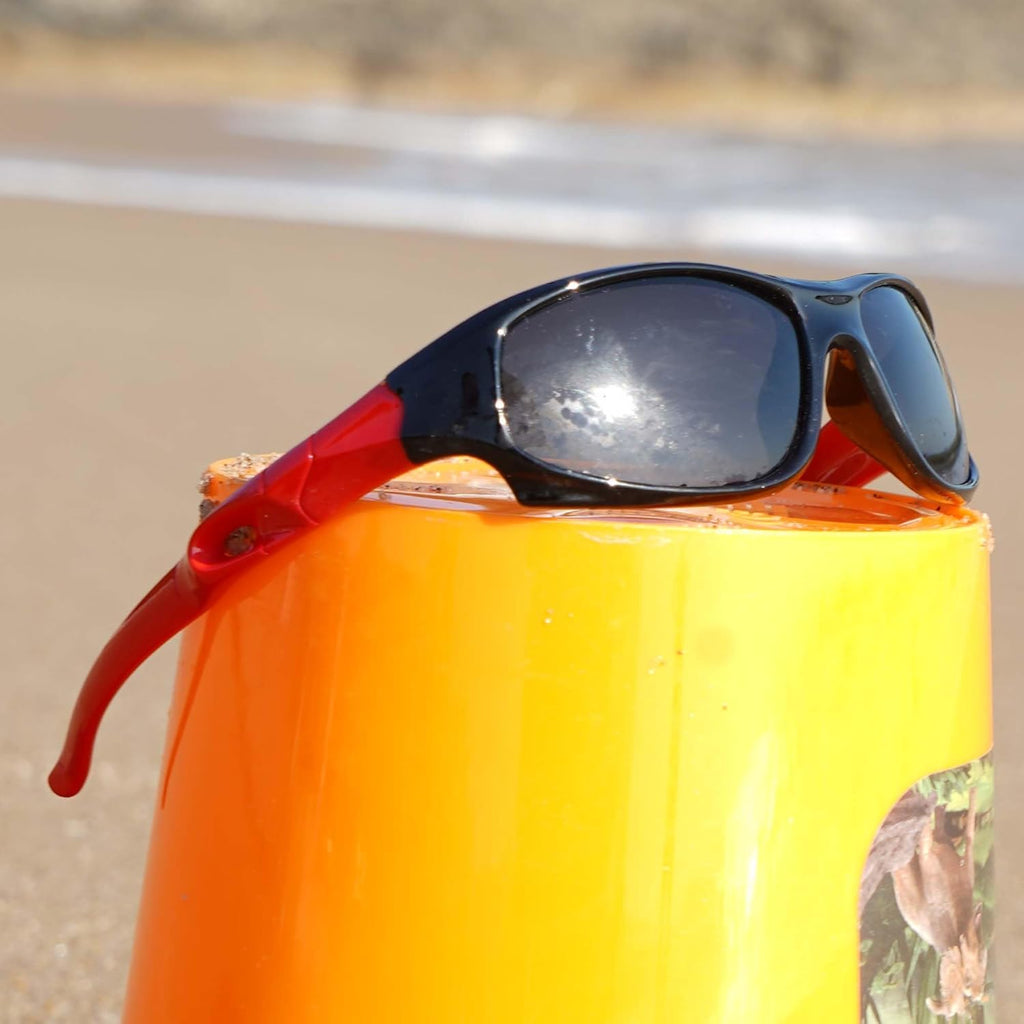 Kids polarized sports sunglasses with black and red frame resting on orange surface at the beach