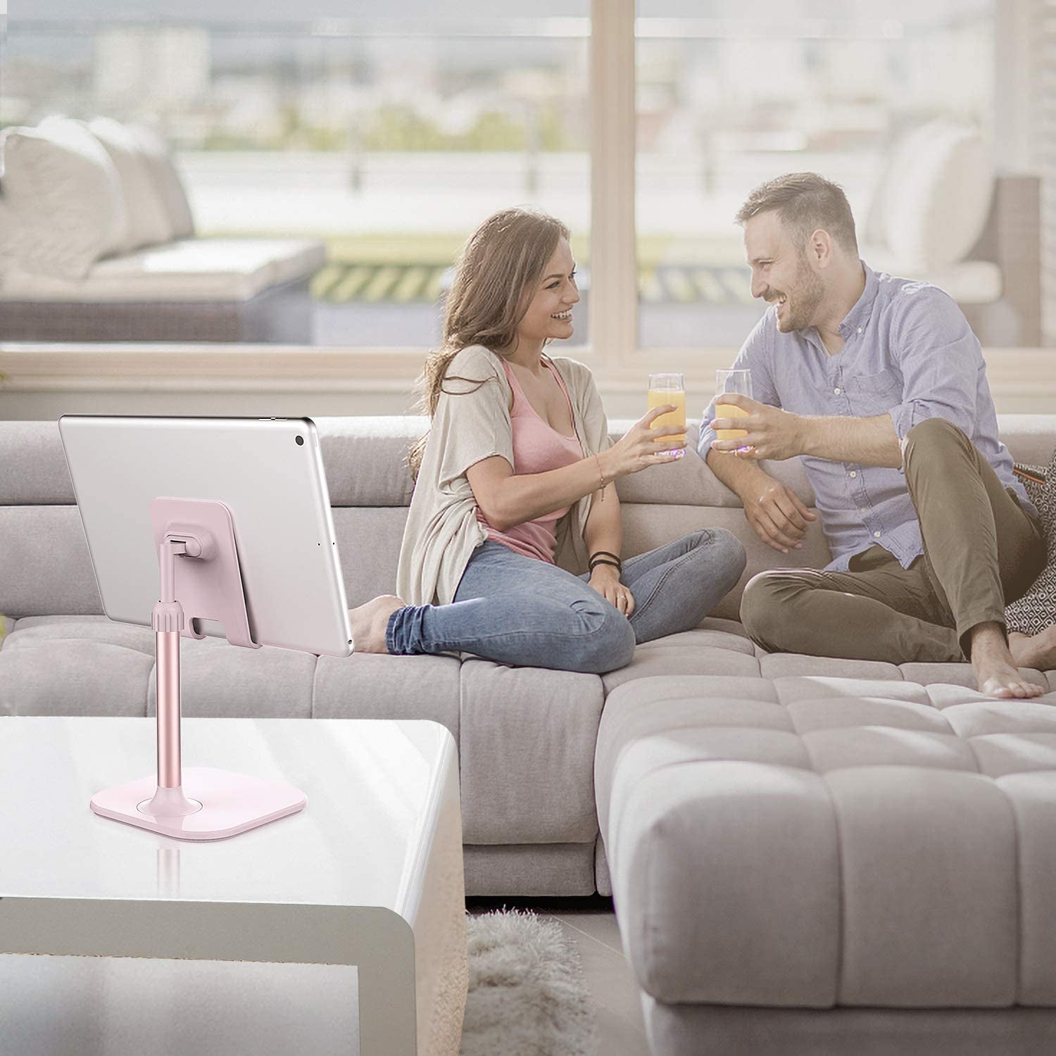 Pink adjustable cell phone stand for desk holding a tablet on white table near couple relaxing on sofa